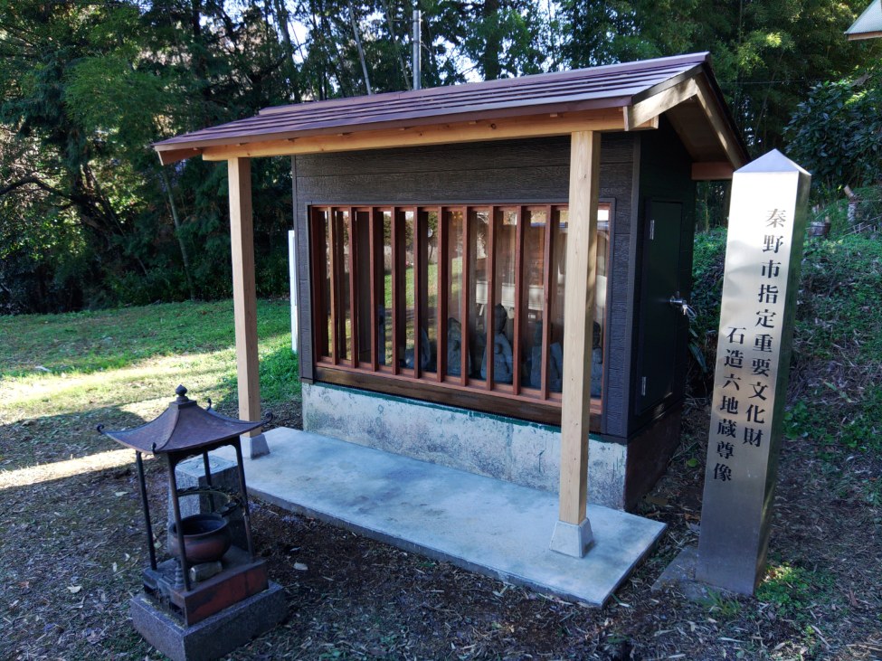 周囲を木々に囲まれ石造六地蔵が安置された小さなお堂が、泰野市指定重要文化財「石造六地蔵尊像」と刻まれた石碑の傍に建っている写真