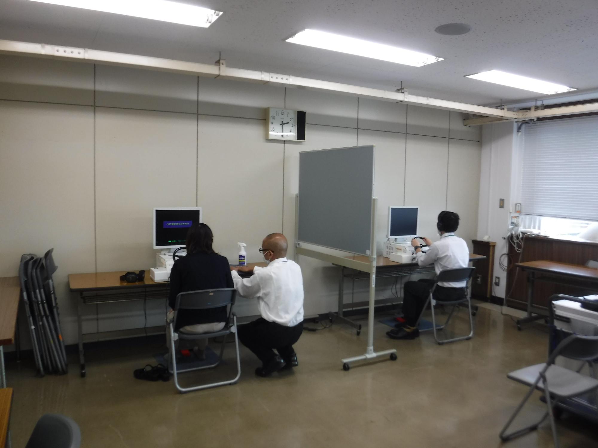 衝立を隔てて並んだ、パソコンが置かれた机に、それぞれ1人ずつが座り、左側の受講者の隣で一人の男性が補助をしている様子の写真