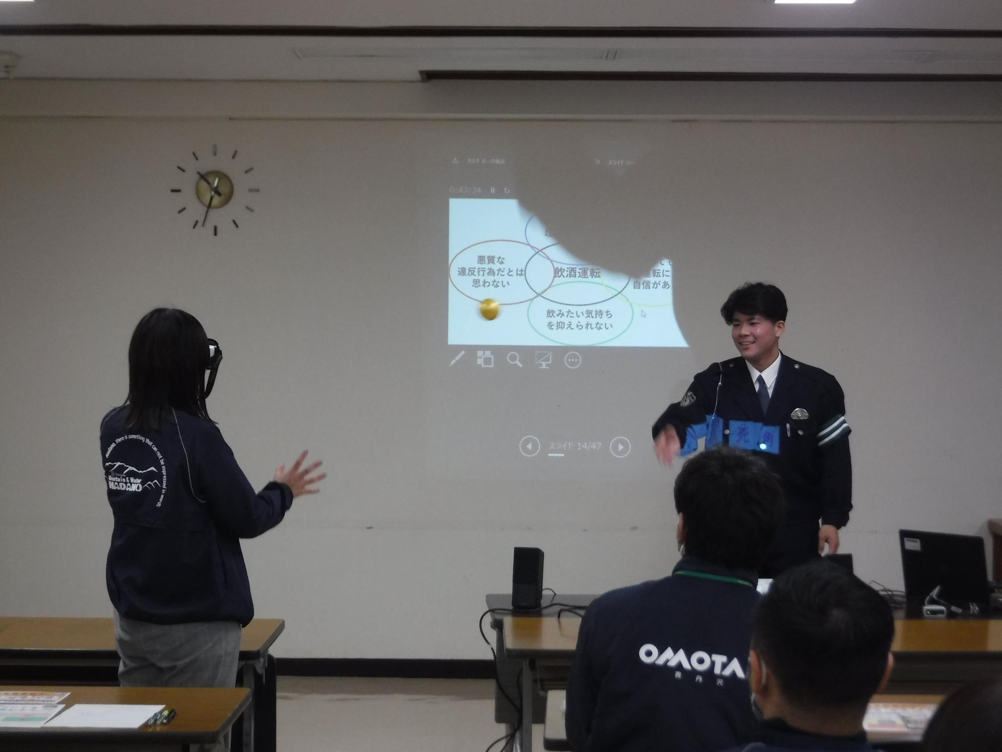 前方の大きなスクリーンに画像が映し出されており、ゴーグルのような装置をつけた女性が席を立ち、スクリーン前に立っている男性警察官の方を向いている写真