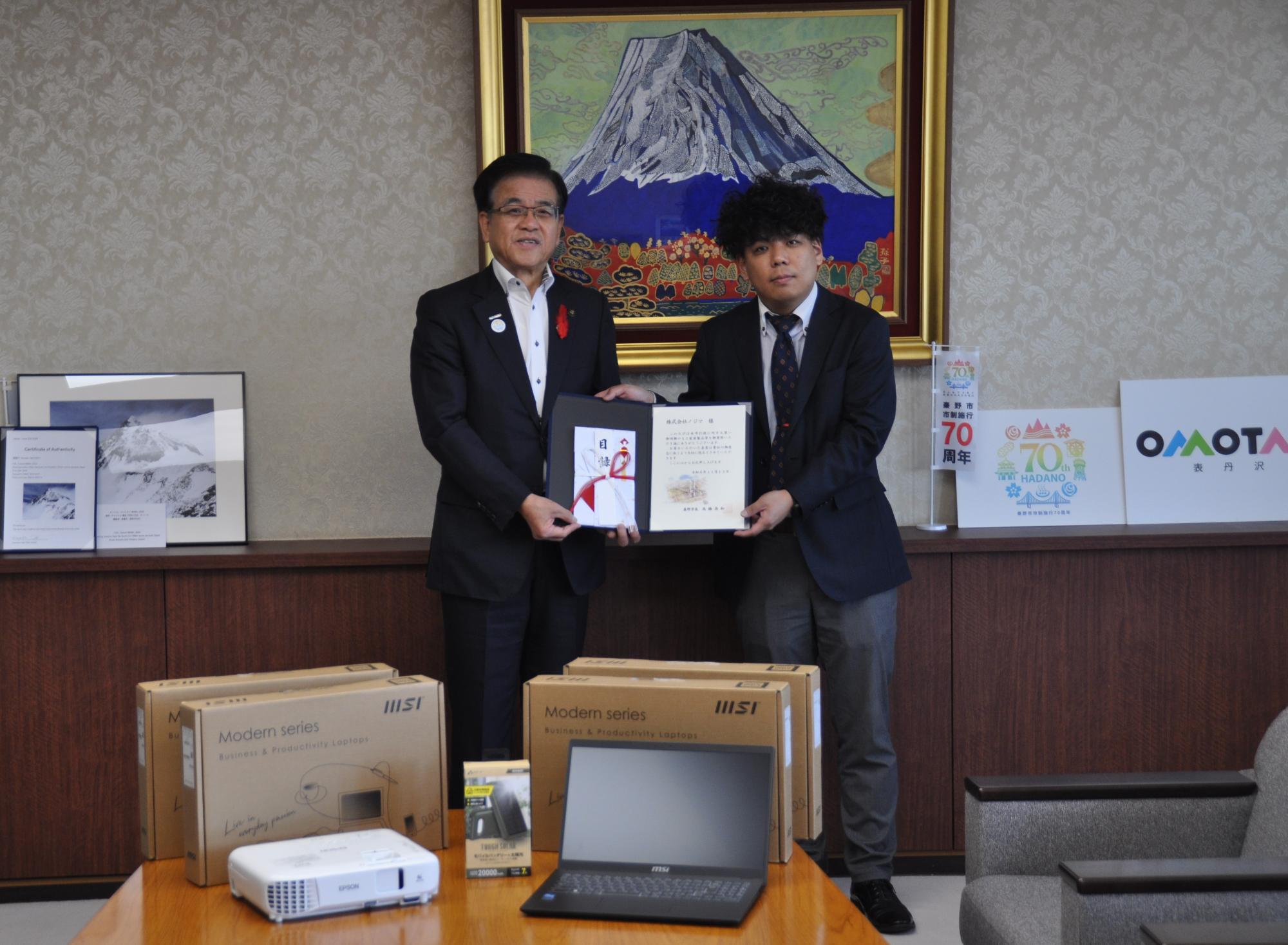 ノートパソコンなどの寄贈品の電子機器が複数置かれたテーブルの後ろで、スーツ姿の市長と男性が感謝状と目録を一緒に持った記念写真