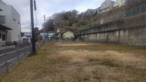 土と枯れ草に覆われた広い更地が広がり、その手前には電柱と道路があり、奥には高いコンクリートの擁壁が見える、住宅地の空き地を捉えた現地写真