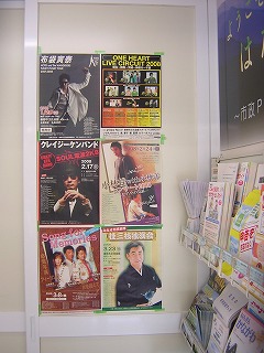 布袋寅泰、クレイジーケンバンド、桂三枝などの音楽や公演のポスターがテープで貼り付けられており、右側には冊子を陳列したラックが見える掲示板の写真
