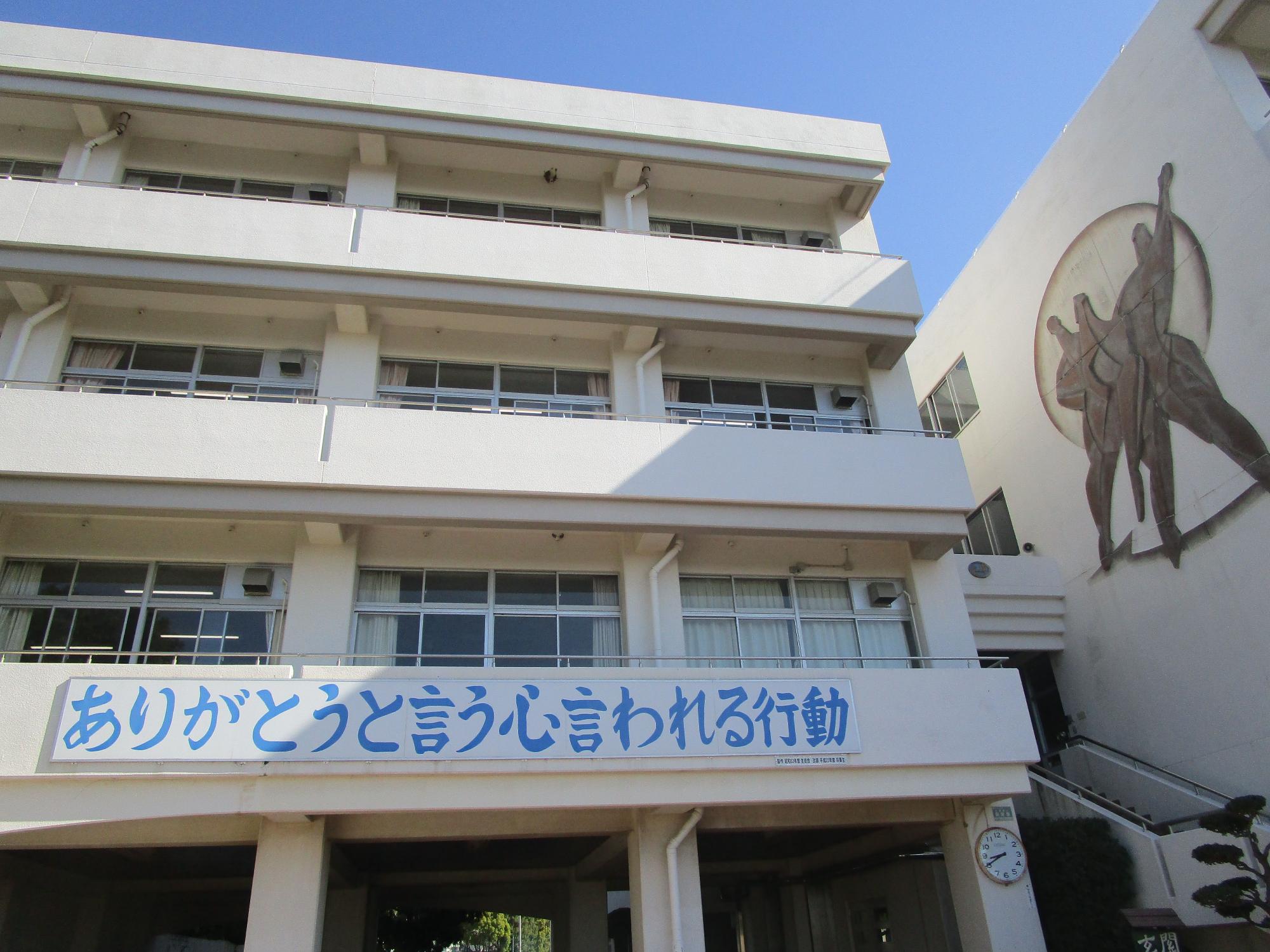 白壁の校舎が写り、中央には「ありがとうと言う心 言われる行動」という青い文字の横断幕のようなものが掲げられ、右側の壁には人物のシルエットのレリーフがある校舎の外観写真