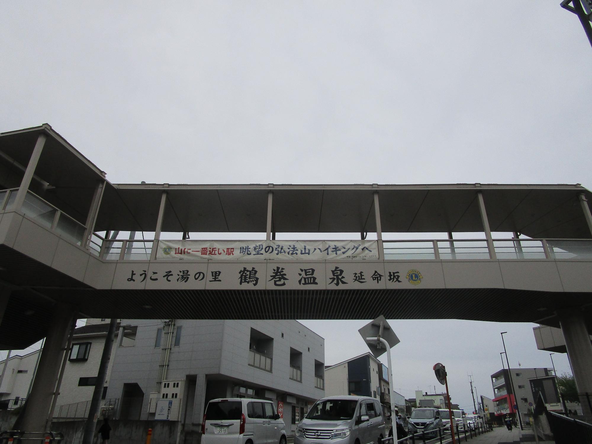 歩道橋の下から見上げる構図で、橋桁の側面に「山に一番近い駅 眺望の弘法山ハイキングへ」「ようこそ湯の里 鶴巻温泉延命坂」の文字が描かれ、その下を複数の自動車が通行している写真
