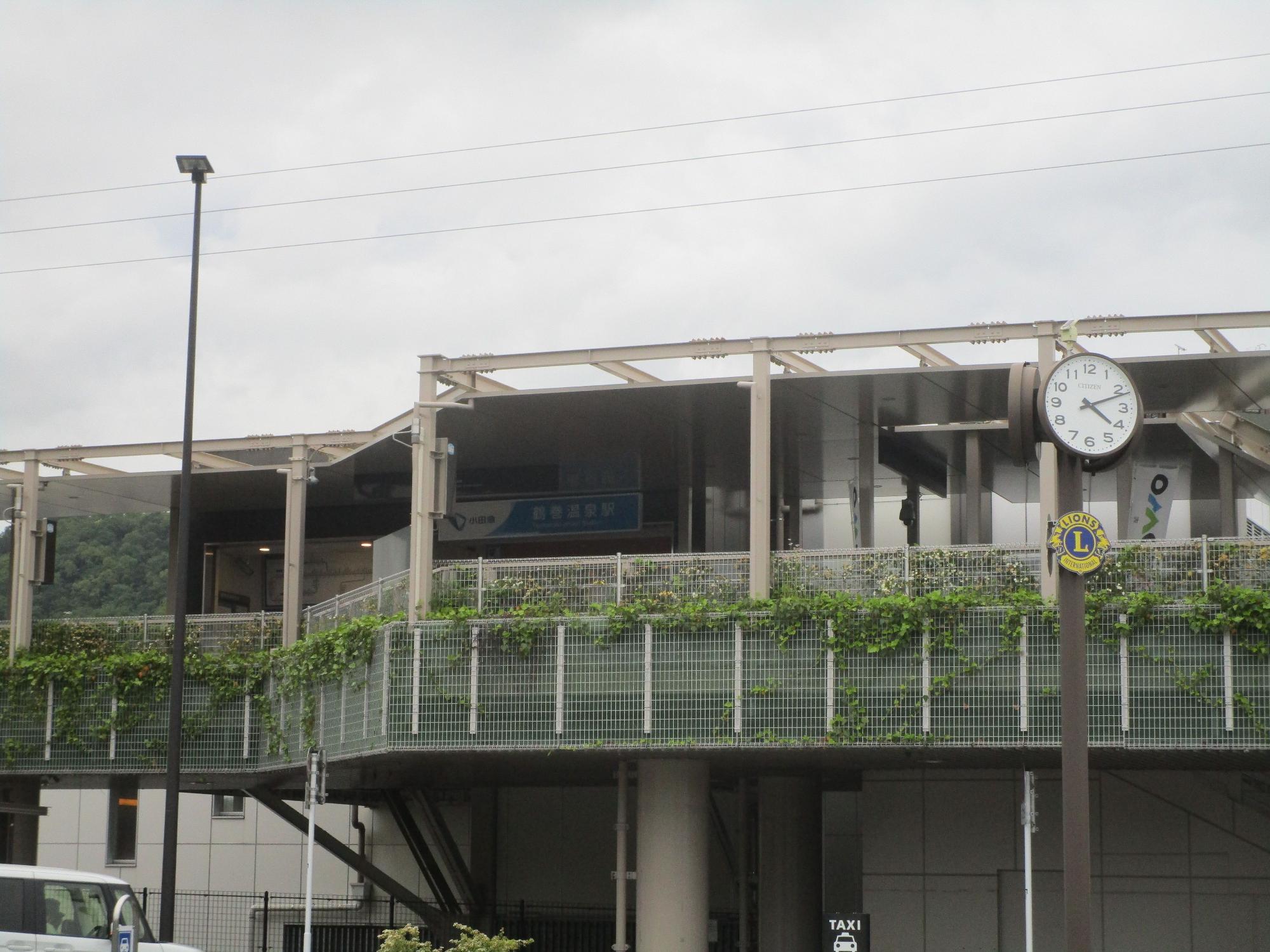 線路の上に架けられた近代的な橋上駅舎の建物で、外側の手すりには緑色のつる性植物が絡みつき、ポールには丸い時計が設置された、曇り空の下の駅舎の外観写真