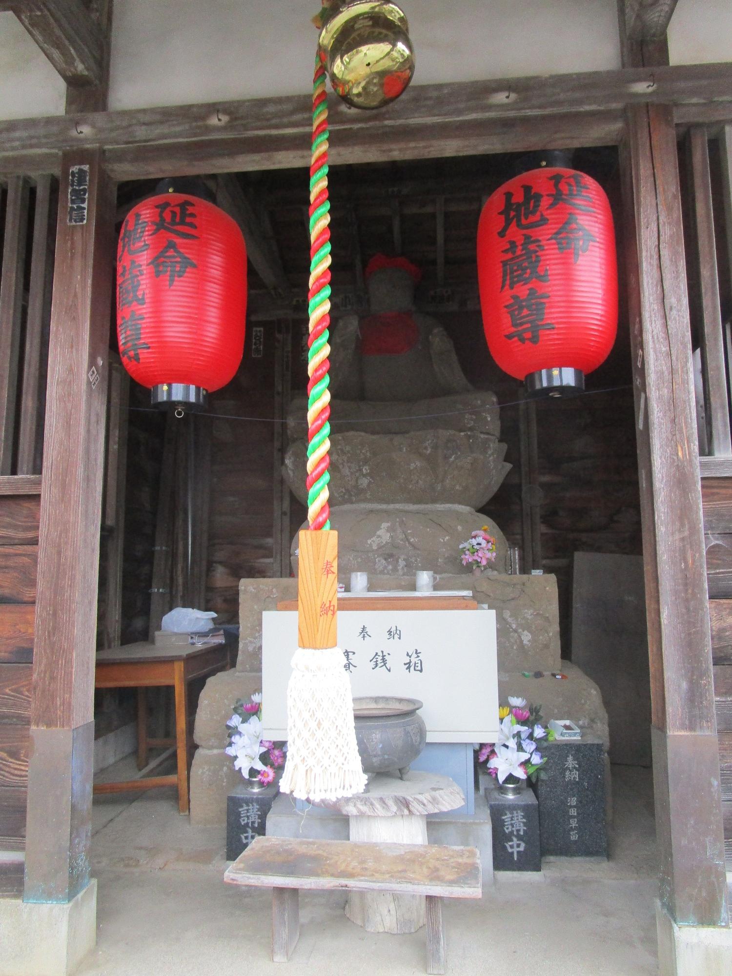 木造の祠の中に、赤い頭巾とよだれかけをつけた石造の地蔵菩薩が鎮座し、その前には奉納賽銭箱と鈴紐、そして左右に「延命地蔵尊」と黒い文字で書かれた赤い提灯が吊り下げられている写真