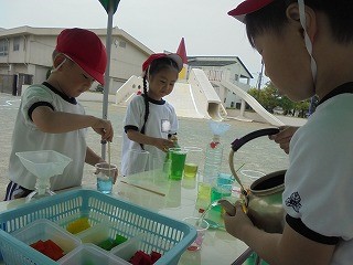 運動帽をかぶった三人の園児たちが屋外のテーブルでやかんや漏斗を使い、水に色をつけたり移し替えたりしている様子の写真