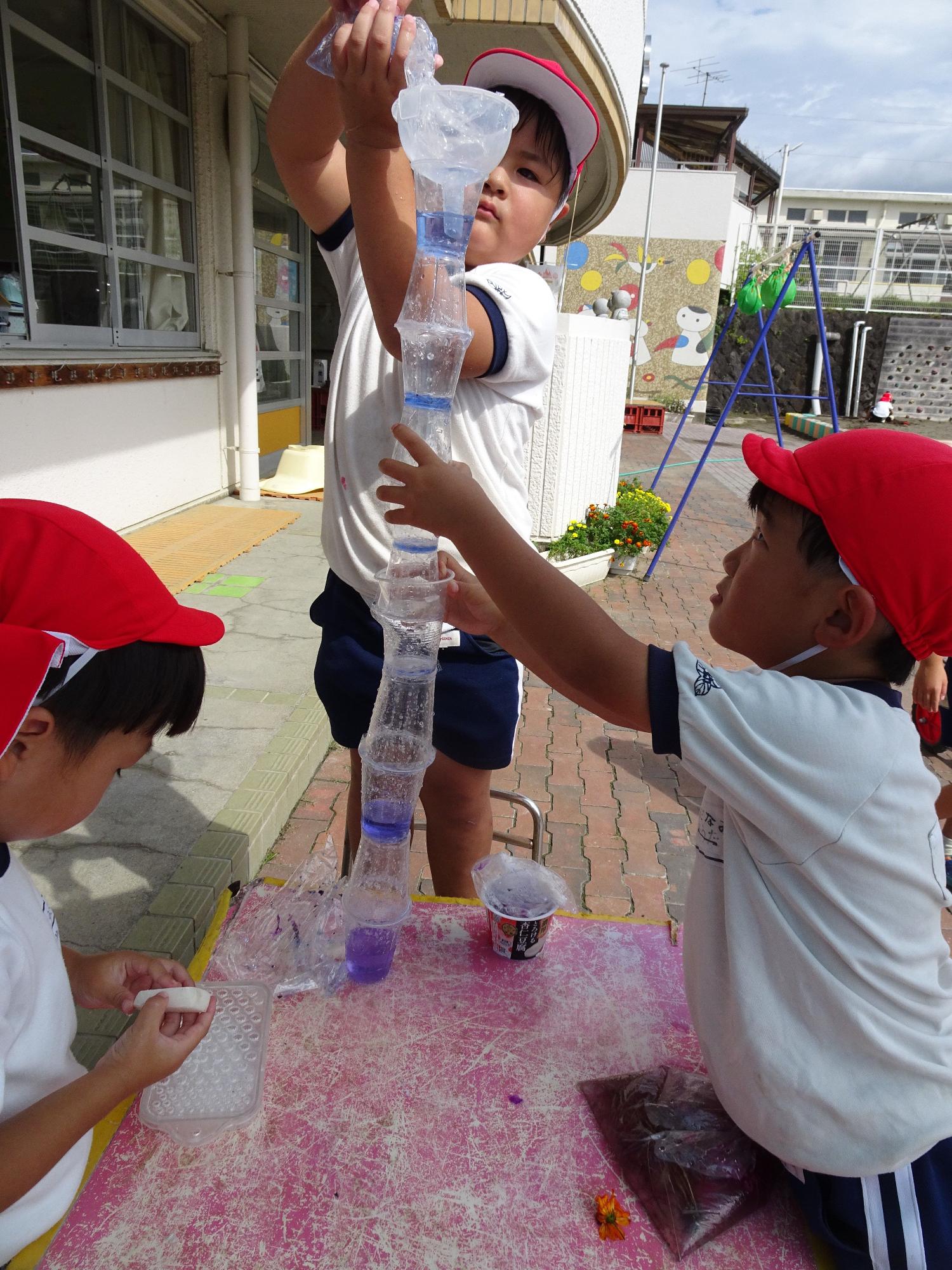 赤い帽子をかぶった三人の園児たちが屋外のテーブルで、積み上げたプラスチックカップを持つ園児と漏斗で色水を注いでいる園児の写真