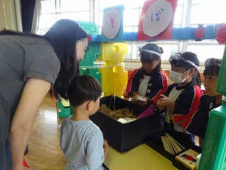 幼稚園の室内で鉢巻を締め法被を着た園児が保護者や他の園児に向かい、そばと書かれた模擬店で紙でできたそばや具材を入れ、そばを売っているごっこ遊びの写真