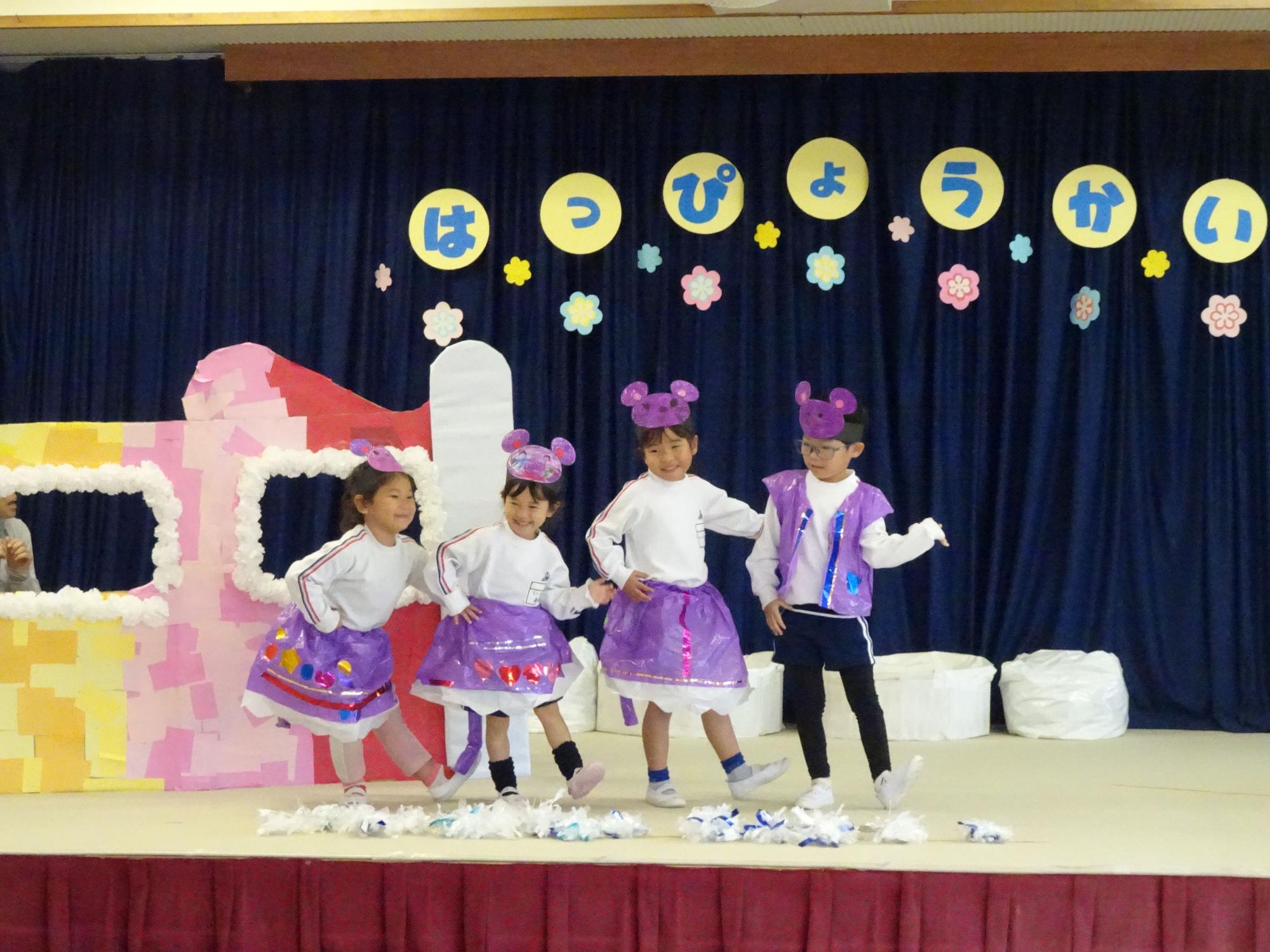 舞台の上で紺色の幕を背景に「はっぴょうかい」の文字と花を飾り付けた中で、紫色のお揃いの衣装を身に着け、紫色のネズミのお面を被り笑顔でダンスを披露する四人の園児たちの舞台写真