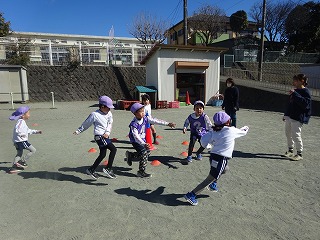 青空の下、広々とした園庭で紫色の帽子をかぶった複数の園児たちが二人の保育士に見守られながらコーンの間を走り回って体を動かす戸外遊びの写真