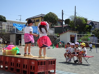 屋外の園庭に設置された台の上で、ピンクのチュールスカートを身につけた女の子2 人が踊っている様子で、その前でベンチに座って見学している4人の園児の写真