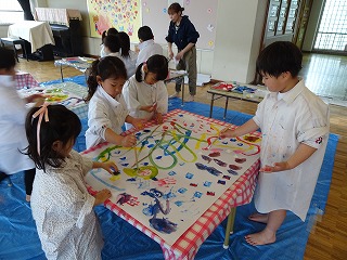 広い室内の床に敷かれた青いシートの上、白いシャツを着た複数の園児たちがテーブルに置かれた大きな白い紙に色鮮やかな絵の具で絵を描いている写真