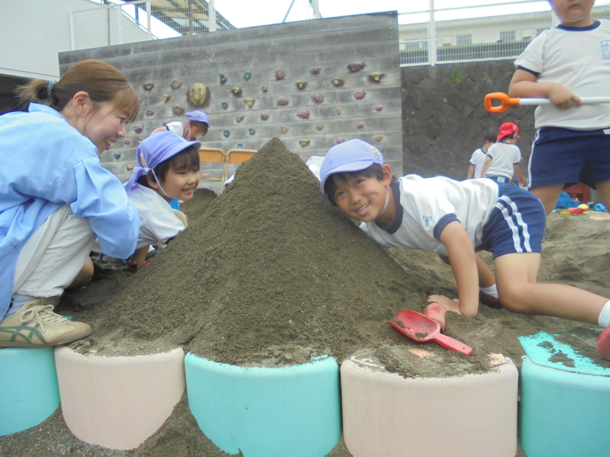 砂場で大きな山にトンネルを作り笑顔で顔を覗かせる水色の帽子をかぶった二人の園児と、しゃがんで優しく見守る先生の写真