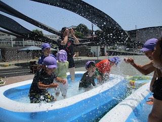 屋外で、紫色の帽子をかぶった園児たちがビニールプールに入り、はしゃいで水をかけ合っている写真