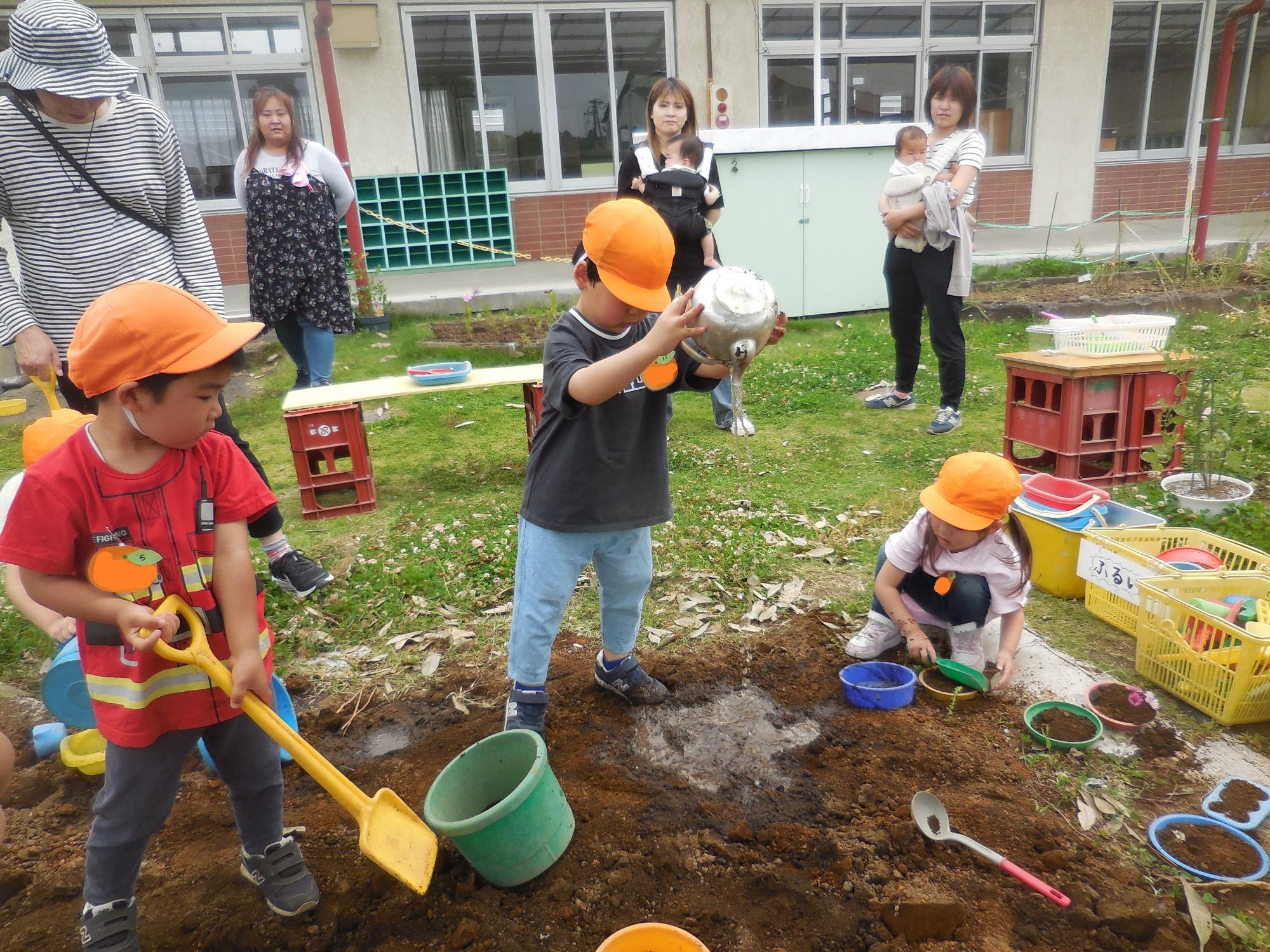 オレンジ色の帽子をかぶった三人の園児が、庭の土の上でスコップやバケツを使って泥遊びに夢中になっており、その後ろから複数の大人が見守っている写真