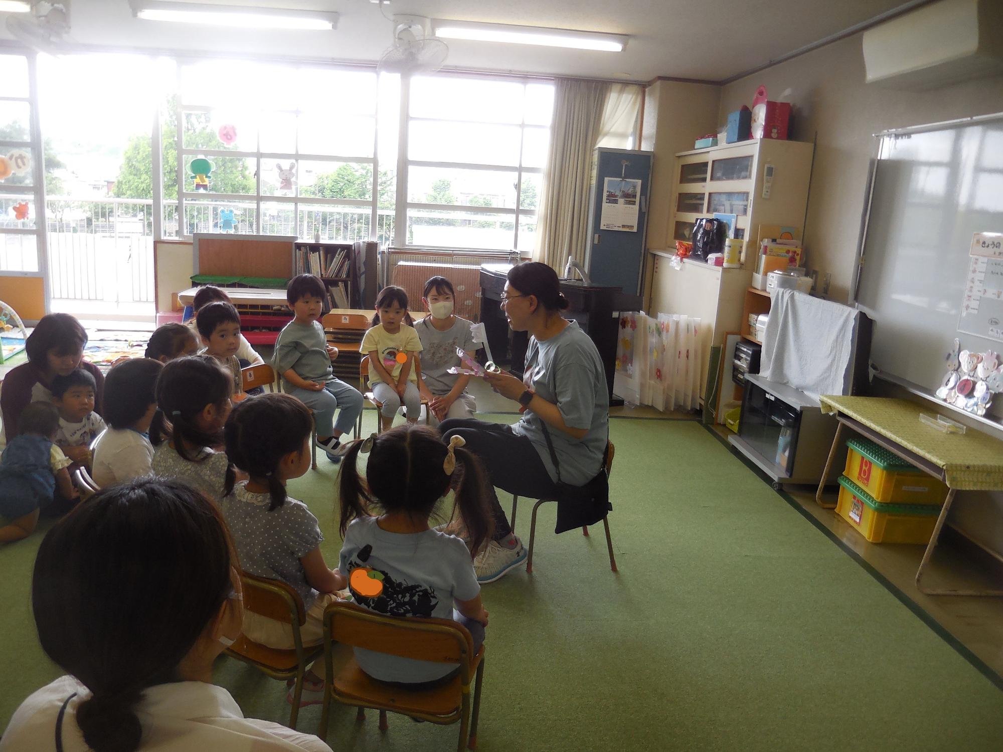 明るい窓のある広々とした教室で、一人の先生が床に敷かれた緑色のカーペットの上で椅子に座り、数十人の園児たちを前に大きな歯ブラシを持ち話をしている様子の写真