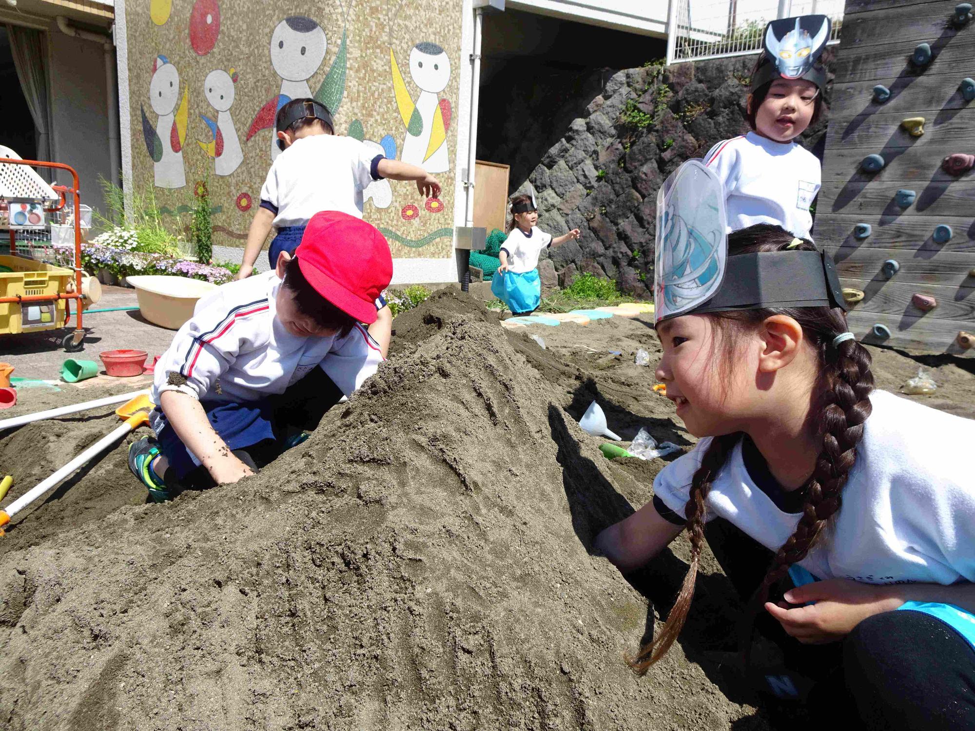 年少児が砂場で山作りをし、2人の子どもが互いに目の前の斜面を掘り進めてトンネルをつなげようとしているところです。