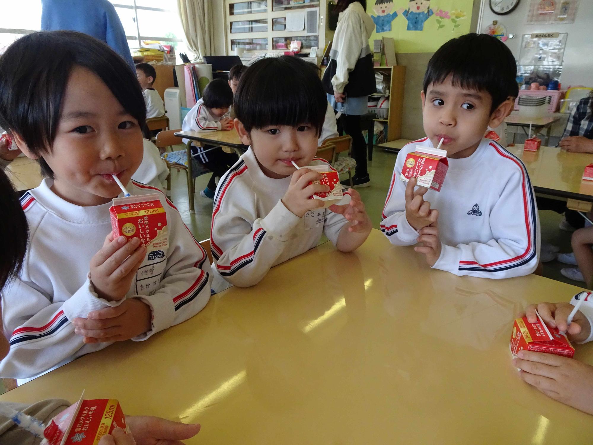 年長児と年少児が椅子に座り、一緒に牛乳を飲みながら休息しているところです。