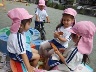 ピンク色の帽子をかぶった3人の園児が園庭の遊具の近くに集まり、座って落ち葉を持ち合遊んでいる様子の写真