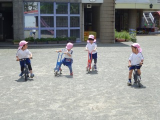 広々とした園庭の地面で、ピンク色の帽子をかぶった四人の園児たちがそれぞれバランスバイクやキックスクーターに乗り、楽しそうに遊んでいる写真