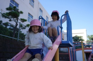 青空の下、幼稚園の園庭でピンク色の帽子をかぶった二人の園児が、楽しそうに笑顔でピンク色の滑り台を滑っている写真