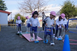 青空の下、ピンクの帽子をかぶった複数の園児たちが、ぽっくりを履いて板で作られた板の上を歩き笑顔で遊んでいる写真