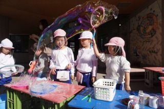 戸外のテーブルで遊ぶピンクと白の帽子をかぶった園児たちが、大きなシャボン玉を作り出し、虹色に輝くシャボン玉を見ている写真