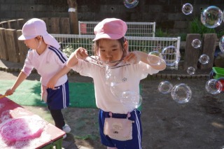 太陽の光が当たる園庭で、ピンク色の帽子をかぶった一人の園児が両手で細長い輪を持ち、たくさんのシャボン玉を作っている写真