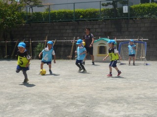 園庭で青い帽子と水色のビブスと黄色のビブスを着用した5人の園児たちが、指導員の男性に見守られながらゴールに向かってサッカーを楽しんでいる写真