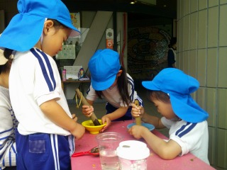 青い帽子と体操服を着た三人の園児たちが、テーブルの上でボウルに入った水などを木の棒で熱心にかき混ぜて遊んでいる写真