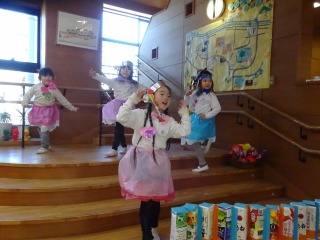 木目調の階段のある室内で、手作りの衣装を着て頭飾りをつけた四人の園児が、楽しそうにダンスを披露している写真