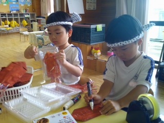 ねじり鉢巻きをつけた体操服姿の二人の園児が、室内で茶色の折り紙に書いたり、透明のプラスチック容器に茶色の折り紙で作った食べ物を梱包している、模擬店のごっこ遊びの準備をしている工作風景の写真