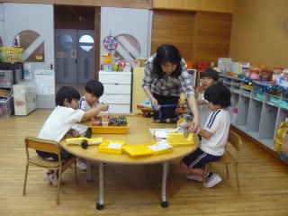 教室で先生が円卓で4人の幼児と一緒にブロックや型はめのような教材を使い、楽しそうに遊んでいる様子の写真