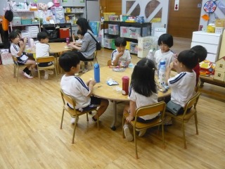 木の床と明るい照明の教室で、2つの円卓に分かれて複数の幼児が座り、おやつを食べている写真