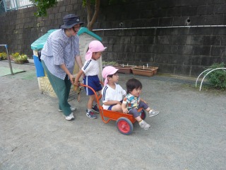 園児と小さな男の子が仲良く座った赤い手押し車を、園児と女性が一緒に押している写真