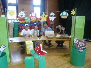 いろいろなキャラクターの飾り付けがされた屋台のような場所で、中央のテーブルを囲んだ4人の方が楽しそうに座っている写真