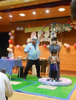 水玉模様のシャツを着た男性が、大きなシャボン玉の輪を使って、台の上に立つ母親と小さな子どもをすっぽり覆う巨大なシャボン玉を作ろうとしている屋内イベントの様子を捉えた写真