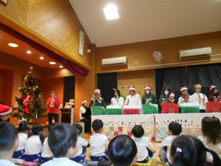 クリスマスツリーとクリスマスの飾り付けがされたホールのような場所で、サンタ帽などをかぶった大人たちが並んでハンドベルのような楽器を演奏し、それを見上げるたくさんの小さな子どもたちが床に座っている様子を写した写真