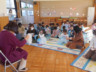 フローリングの床に敷かれたマットの上に座った多くの親子たちが、水色や白の紙のような素材で制作活動に取り組んでいる様子の写真