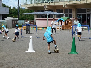 白や緑色のコーンやゴールが設置された広い園庭で、数人の先生が見ているなか、水色の帽子を被った子供たちがサッカーの練習をしている様子の写真