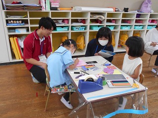 幼稚園の教室で二人の園児がテーブルで製作活動をしており、マスクをした保育士と赤いポロシャツの男性が見守っている写真