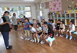 幼稚園の明るい教室で、一人の先生が絵本を手に持ち、椅子に座った十数名の園児たちに読み聞かせをしている写真