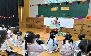 教室で、一人の先生が前に座ってカボチャの紙芝居を読んでいるのを床に座った複数の園児が話を聞いているのを後方から写した写真