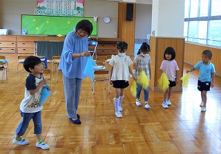 広い室内で一人の保育士が、黄色や水色のシフォン布のようなものを持って集まった五人の園児たちと遊びを楽しんでいる様子の写真