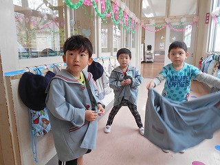 上着を着た三人の園児たちが、制服が掛けられた廊下の前で立ち止まり、制服を着脱している写真