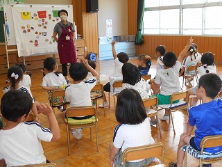 広々とした室内で、一人の保育士がホワイトボードに貼られた食べ物の絵を見せながら、椅子に座った十数名の園児たちに質問をして、園児たちが元気に手を挙げている写真