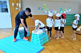 体育館のような室内で、体操服と帽子姿の園児たちが体育指導員の男性に見守られ、青と白の跳び箱をしている写真