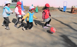 戸外の広場で赤い帽子と赤のビブス、白い帽子と水色のビブスを着用している4人の園児が赤いボールを蹴りながら追いかけてサッカーをしている写真