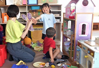 室内のプレイエリアで、女性が膝をついておもちゃの羽を笑顔の女の子に渡し、男の子も一緒に大きなドールハウスの前に座りおままごとをしている写真